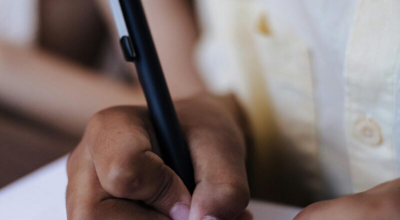 Child's hands writing with a pen on paper indoors, showing focus and concentration.